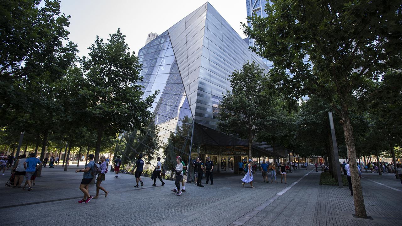  Dozens of people walk in the shade of oak trees outside the Museum Pavilion on a warm, sunny day.